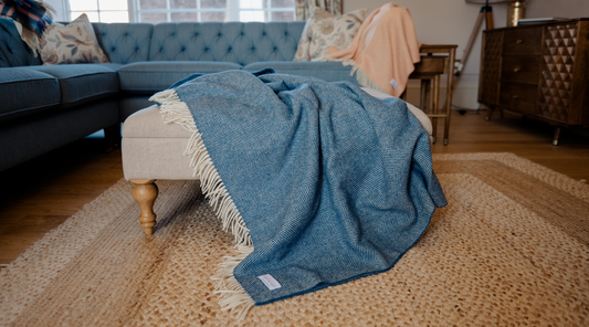 Blue herringbone wool blanket with cream fringe draped over an ottoman in a cosy living room – classic British style meets modern interior design.