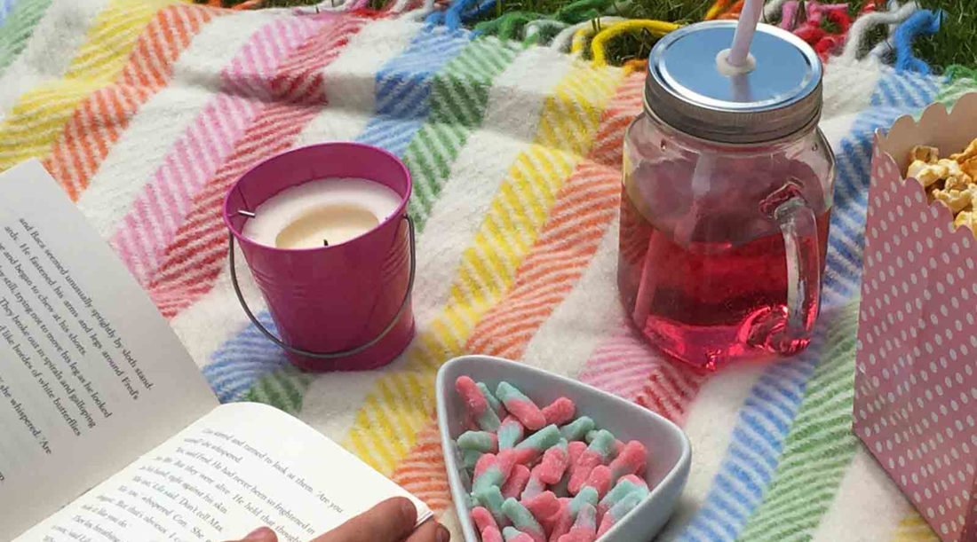 Picnic blanket laid out in the sun with person reading, drink and snacks