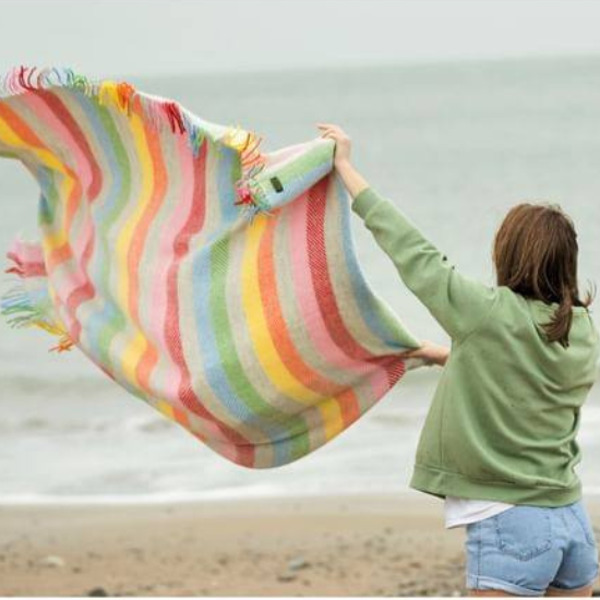 Woman shaking out a colourful striped wool blanket on a breezy beach – perfect for outdoor adventures, coastal picnics and cosy camping moments.