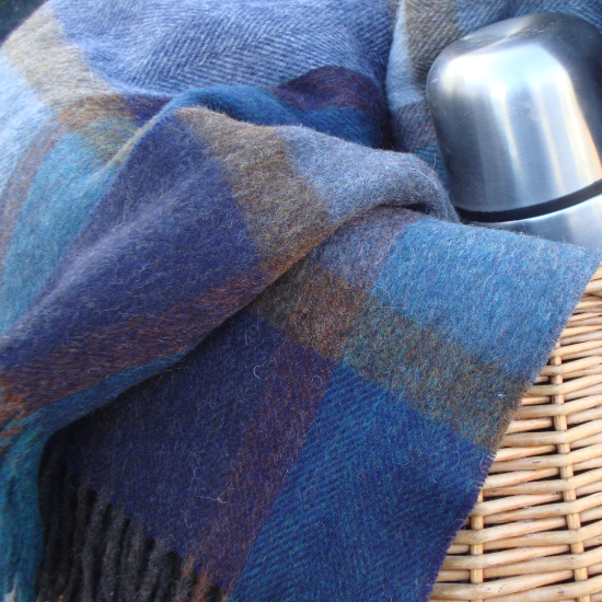 Close-up of a rich blue check lambswool throw draped over a wicker picnic basket with a stainless steel flask – part of the Solway Blankets Blue Throws Collection.