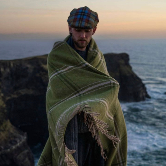 Man wrapped in a traditional green wool blanket with fringe, standing on dramatic coastal cliffs at sunset – a rugged and thoughtful gift idea for him from Solway Blankets.