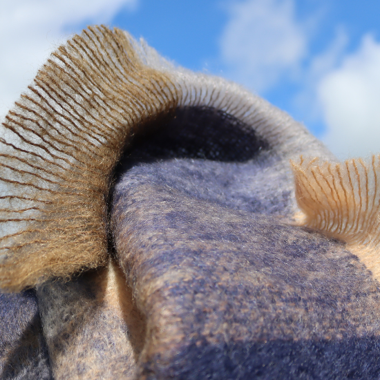 Close-up of a soft mohair throw in natural blue and tan tones, gently blowing in the breeze against a blue sky – lightweight warmth with luxurious texture.