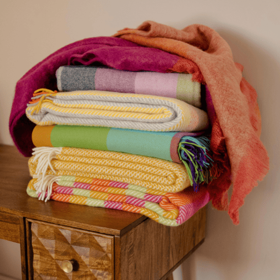 Stack of colourful wool blankets and throws in vibrant checks, stripes, and solids – a cheerful mix of British-made designs perfect for brightening your home.