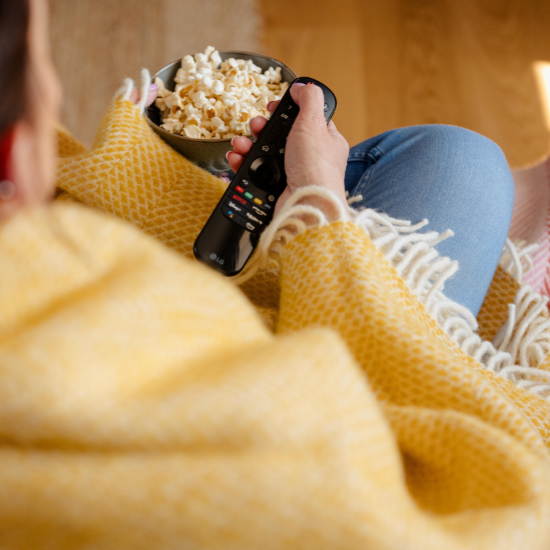 Person relaxing under a mustard yellow wool throw with twisted fringe, holding a remote and enjoying popcorn – cosy home scene from the Solway Blankets Mustard and Yellow Collection.
