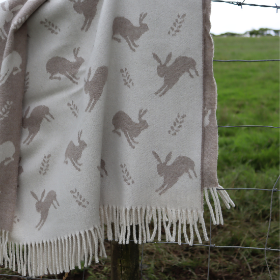 Reversible wool throw with a charming hare and wheat motif in soft taupe and cream, draped over a fence post in the countryside – from the Solway Blankets Reversible Collection.