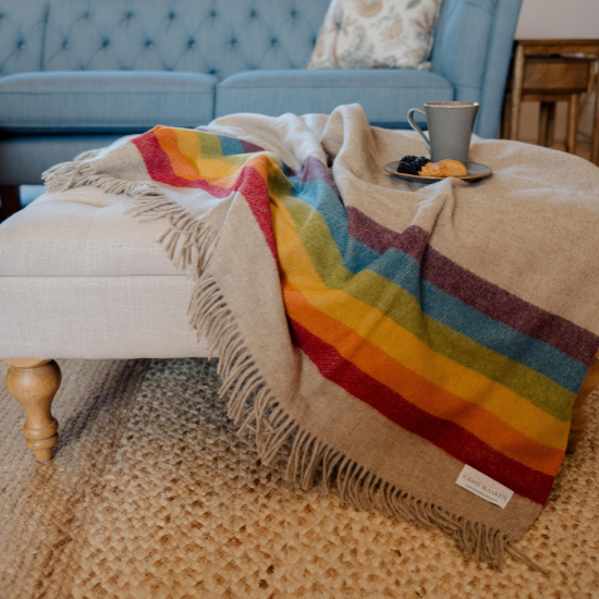 Cosy neutral wool throw with bold rainbow stripes draped over a footstool in a stylish living room – part of the Stripe and Spot Blankets and Throws Collection by Solway Blankets.