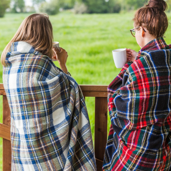 Two women wrapped in cosy tartan wool travel rugs enjoying a warm drink outdoors – perfect for countryside getaways, picnics and year-round adventures.