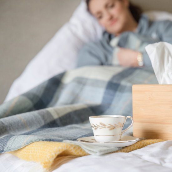Woman resting under a soft checked wool blanket in bed, with a cup of tea and tissue box nearby – part of the Solway Blankets Thinking of You Collection, designed to comfort and care.