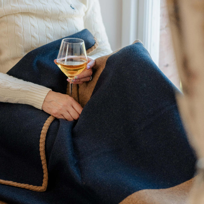 Person sitting by a window holding a glass of wine, wrapped up in a navy blue and tan blanket.