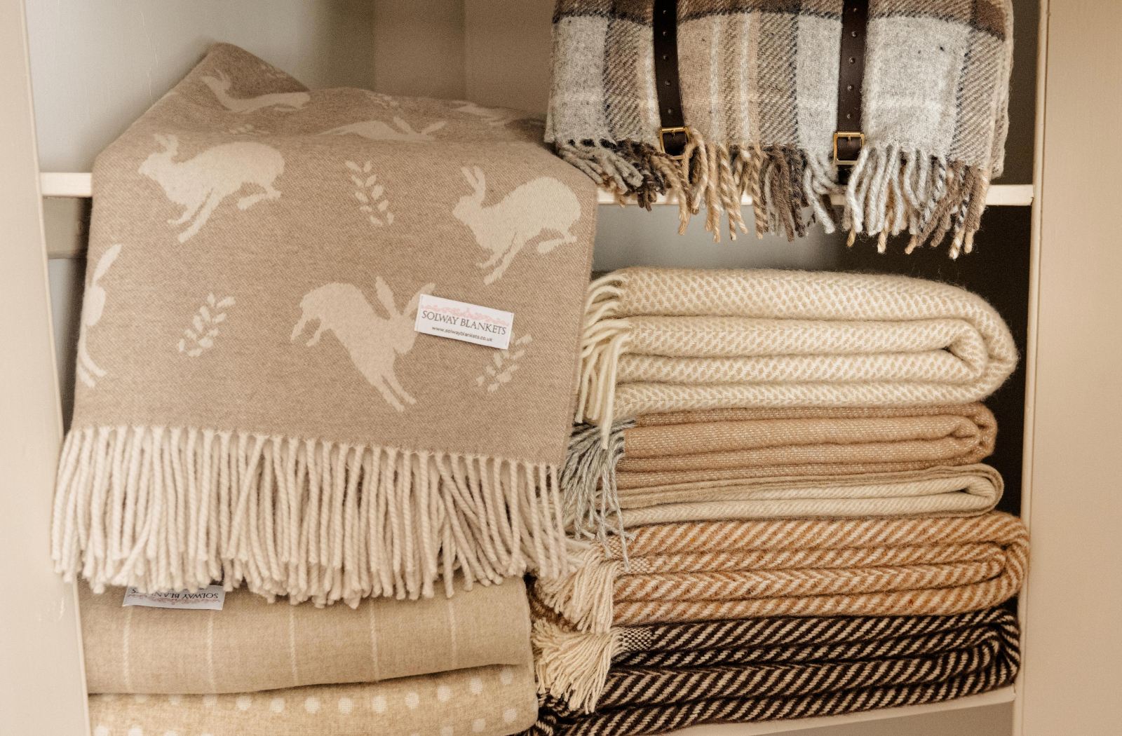 Collection of beige and brown patterned blankets in different types of wool with fringes on a wooden shelf.