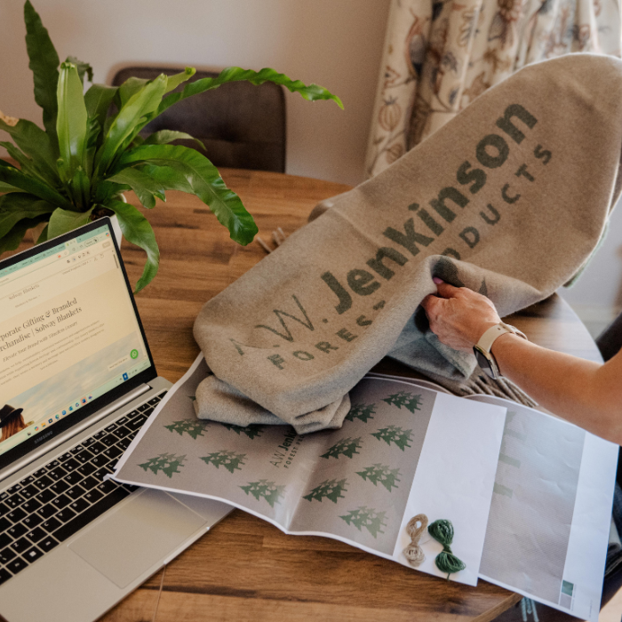 Person holding A W Jenkinson branded bespoke woven blanket, with artwork and yarn samples in foreground and laptop
