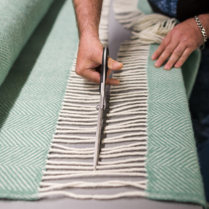 Person cutting a herringbone woven blanket fabric with scissors through the fringe, to create individual finished blankets ready for labelling and folding