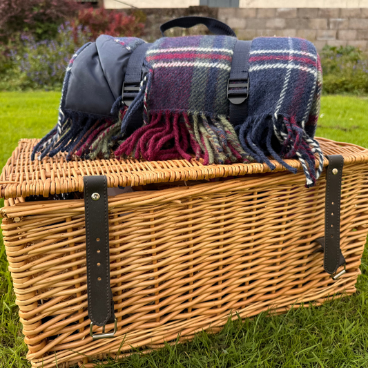 A wicker picnic basket with dark brown leather straps sits on green grass outdoors, with a luxury rolled Morello Cherry tartan picnic blanket with waterproof backing, resting on top.