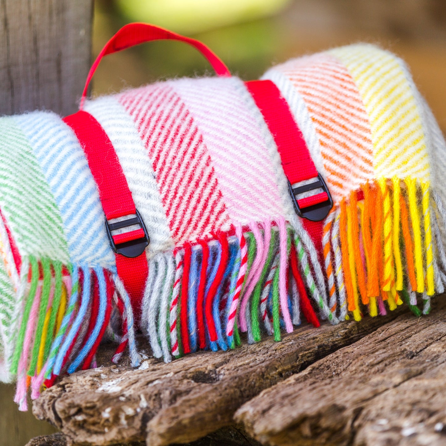 Multicoloured rainbow striped waterproof backed picnic blanket rolled up with red webbing straps, fringes on a wooden surface