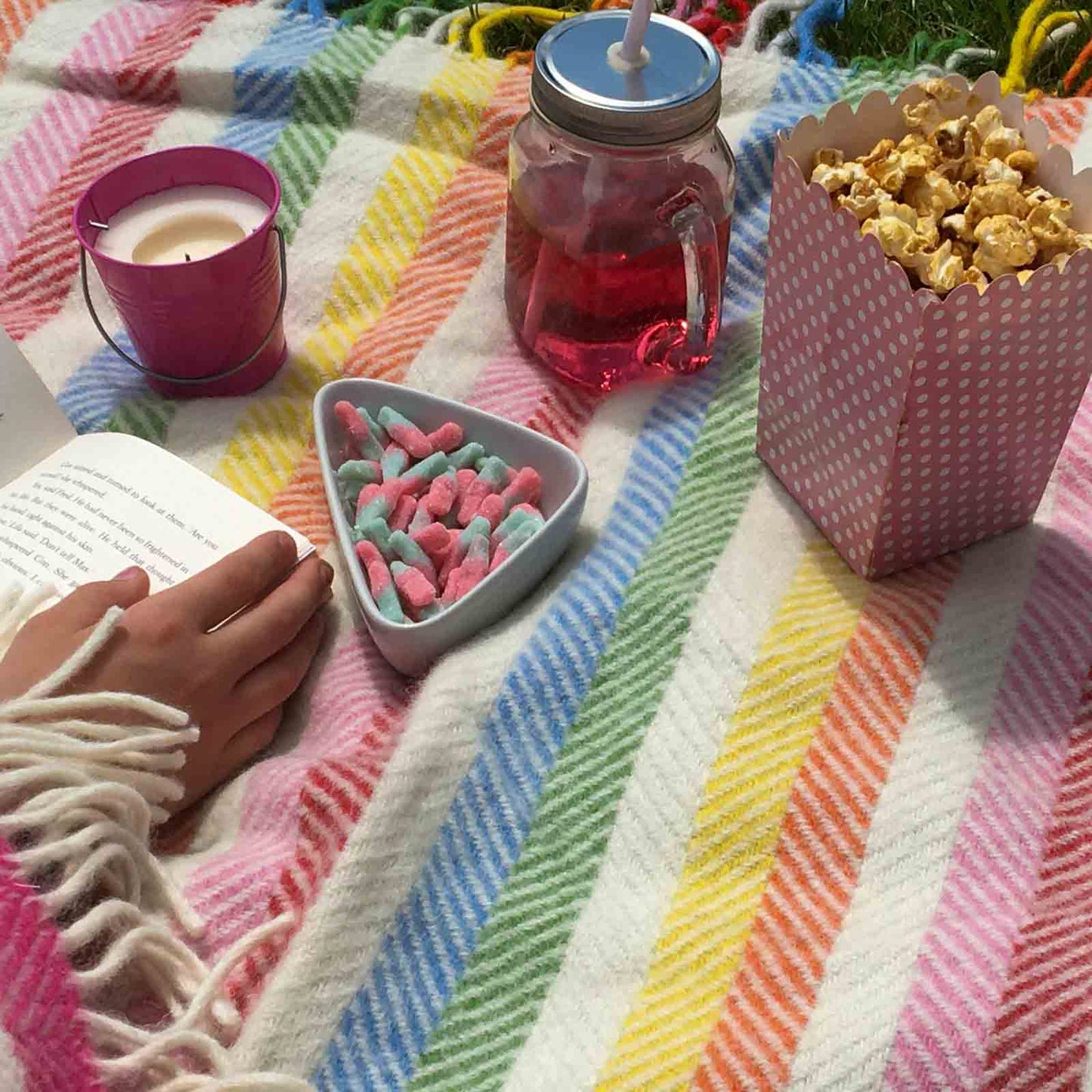 Closeup of pure wool rainbow striped waterproof backed picnic rug with snacks, drink and girl reading