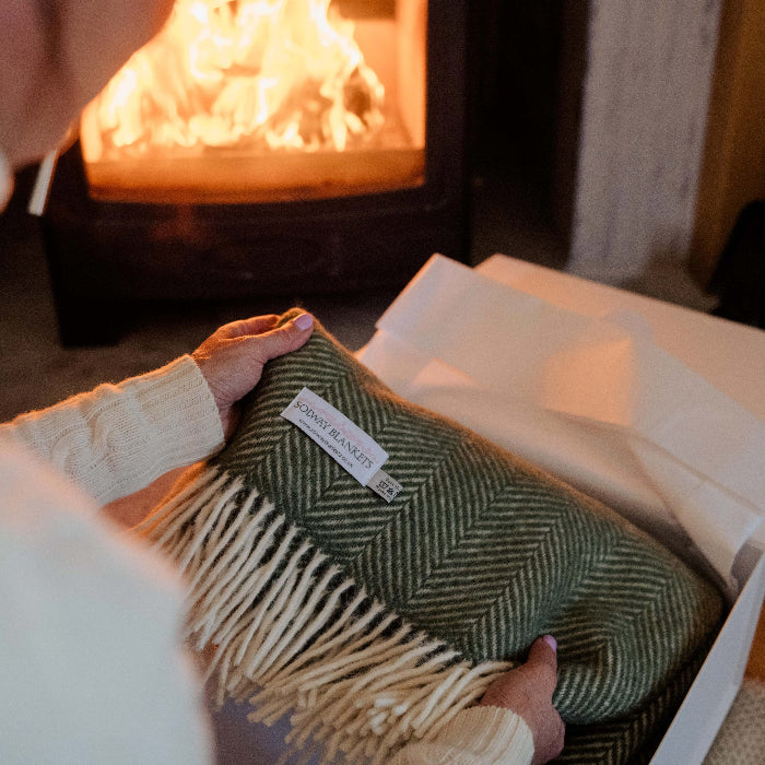 Beautiful Olive Green Herringbone Solway Blankets being gifted to a women in front of the fire, luxuriously wrapped in tissue and presented in a white rigid box with ribbon and gift tag  