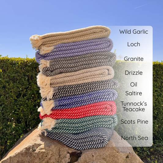 Stack of woven recycled Chevron extra large blankets with colour names on a stone ledge against a blue sky.