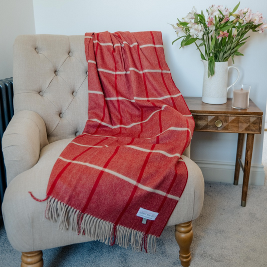 A red and light grey windowpane check cashmere wool throw blanket with fringe is draped over a beige tufted armchair in a styled room setting next to a wooden side table with a vase of pink flowers.