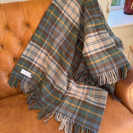 A close-up of the Antique Dress Gordon plaid wool blanket with fringe, featuring muted toned green, blue, brown, off white and yellow checks. A white woven label reading 'SOLWAY BLANKETS' 