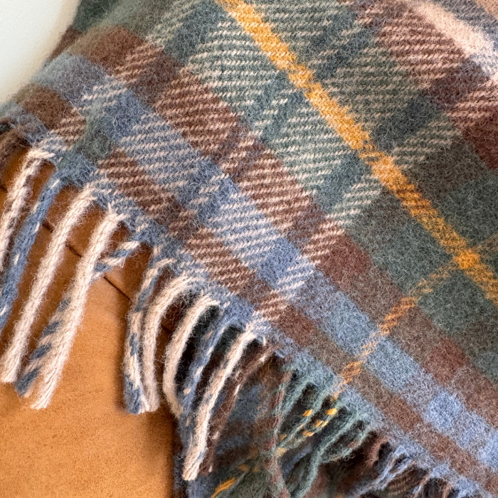 A close-up of a woven Antique Dress Gordon Tartan plaid blanket draped over a leather sofa, showing a multicolour tartan pattern with muted green, brown, blue, off white and yellow, finished with fringed edge