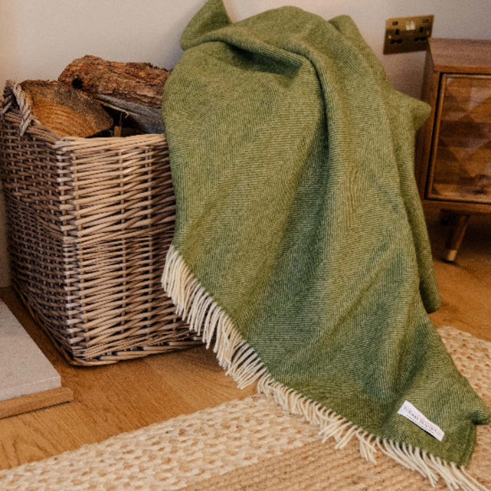 Apple Green Herringbone Shetland Wool Blanket with fringe draped over a wooden basket of next to a wicker basket of logs in a cosy living room