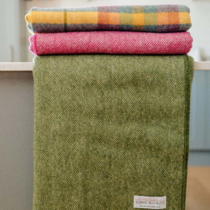 Stack of colourful blankets on a surface with a Apple Green Herringbone Shetland Wool Blanket in the foreground.
