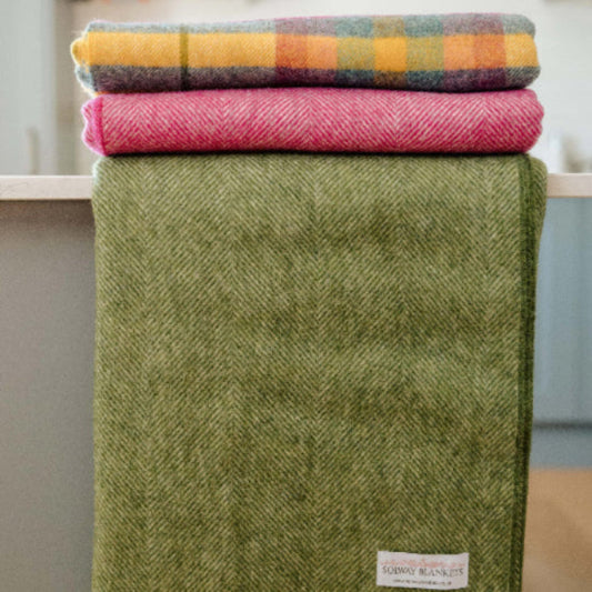 Stack of colourful blankets on a surface with a Apple Green Herringbone Shetland Wool Blanket in the foreground.