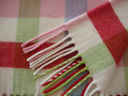 A close-up of a woven bold check baby blanket, Baby Bunting with fringe, featuring a pattern of red, pink, green and cream blocks with a herringbone textured design