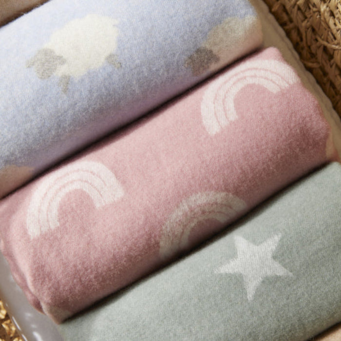 Three folded merino lambswool baby blankets with star, sheep and rainbow patterns on a textured surface.