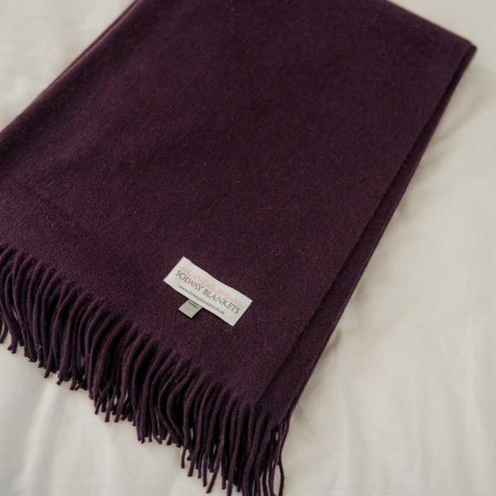 A folded deep plum purple cashmere woven throw blanket with fringe ends and a white label reading 'SOLWAY BLANKETS', laid on a white fabric background.