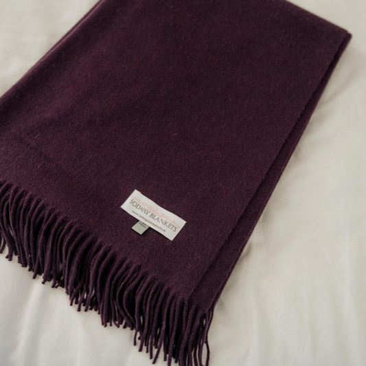 A folded deep plum purple cashmere woven throw blanket with fringe ends and a white label reading 'SOLWAY BLANKETS', laid on a white fabric background.