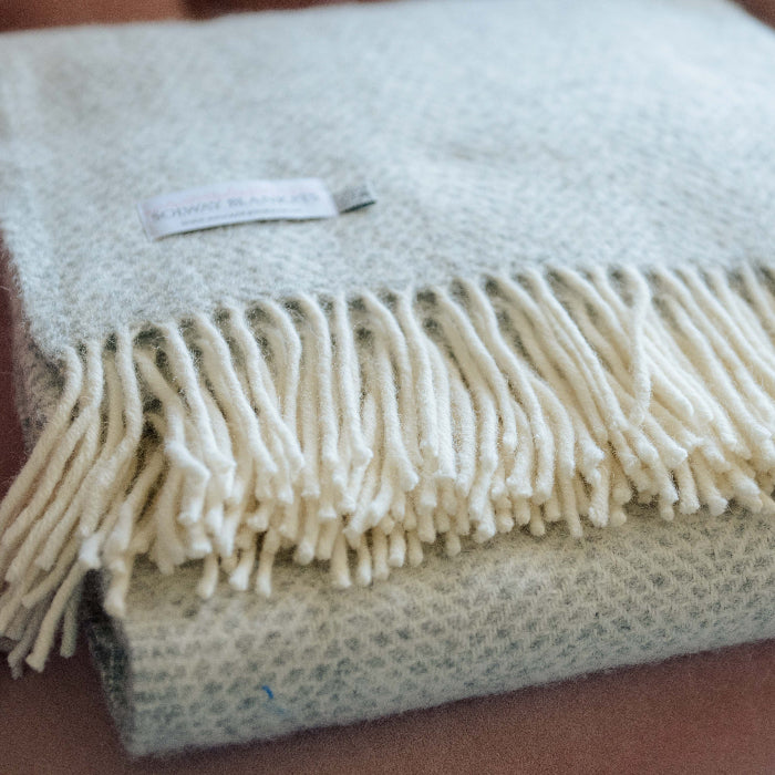 Close up of a grey silver textured wool blanket with white fringe and a Solway Blankets label, folded on a pink chair.