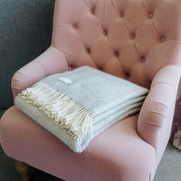 Grey silver textured wool blanket with white fringe folded neatly on a pink armchair, in a home setting.