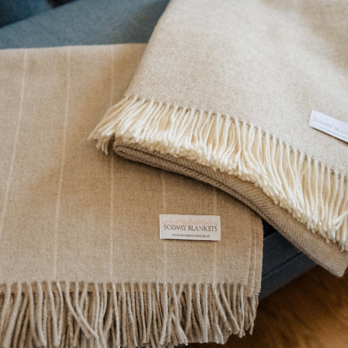 One folded beige pinstripe blanket placed beside a folded beige herringbone plaid blanket with fringes on a blue couch, featuring a visible Solway Blankets label.