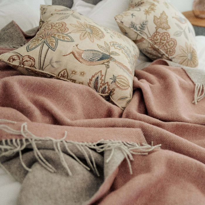 Pink wool throw with a soft grey on the reverse roughly placed on white bedding with decorative and colourful cushions