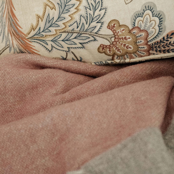 Close up of a pink wool throw with a soft grey colour on the reverse lain infront of a colourful patterned cushion