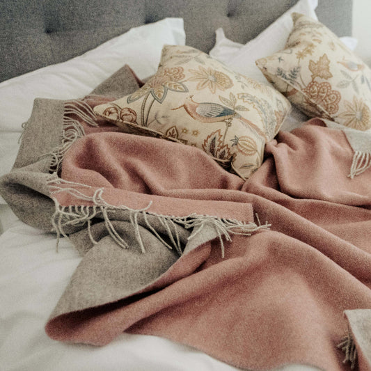 Cosy bedroom with a pink wool blanket throw with a soft grey colour on the reverse side, and patterned pillows on a bed.