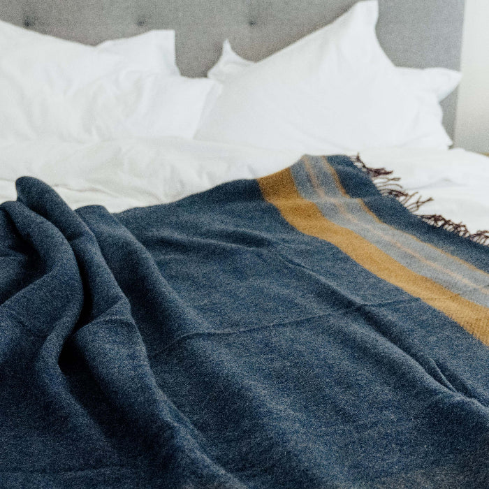 Navy throw with a mustard striped pattern draped over a bed with white pillows and a gray headboard.