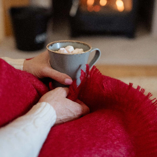 Person holding a mug of hot chocolate with marshmallows in front of a fireplace with the Berry Red Mohair Throw Blanket