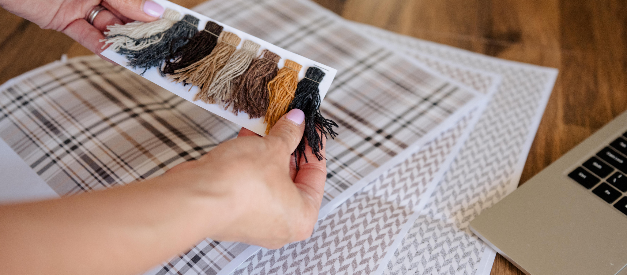 Person holding a tray of coloured yarn swatches against a mock up of blanket designs on paper next to a laptop on a wooden surface