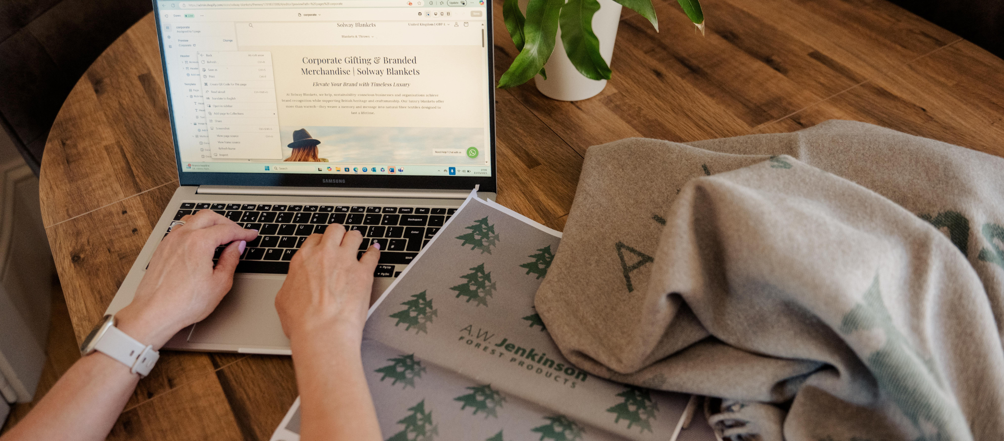 Person using a laptop on a wooden table with a plant, working on a corporate bespoke woven blanket design, artwork design in foreground with customised blanket in the background