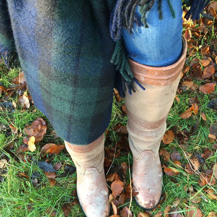 Person wearing brown boots with a green and blue Black Watch Tartan wool knee blanket draped over them, on a grassy background with fallen leaves on the ground
