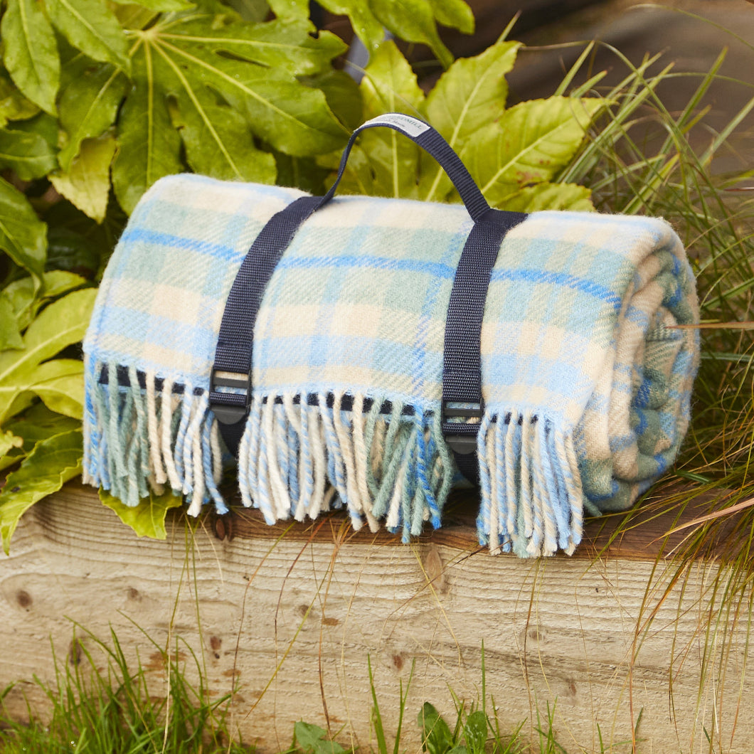 Blue Cottage Plaid waterproof picnic blanket, rolled up with straps and fringe on a wooden bench surrounded by greenery