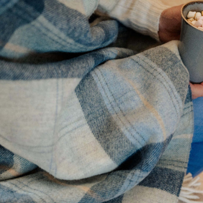 Person wrapped in a Blue Skye Check Shetland Wool Blanket holding a mug with hot chocolate and marshmallows.