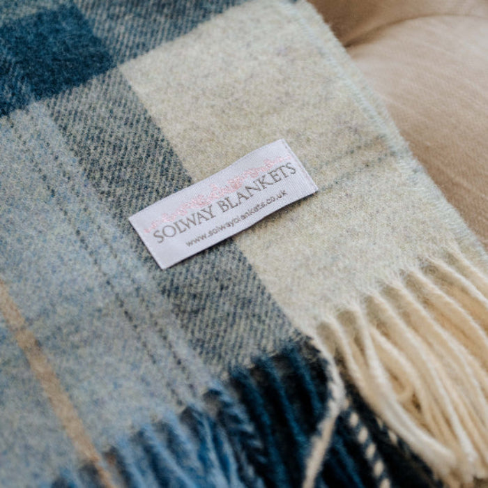 Blue Skye Check Shetland Wool Blanket, navy, duck egg, beige and cream plaid with a visible Solway Blankets brand label on a beige footstool