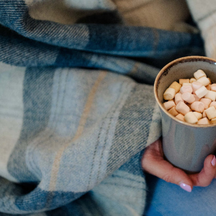 Person holding a mug of hot chocolate with marshmallows, wrapped in a Blue Skye Check Shetland Wool Blanket.
