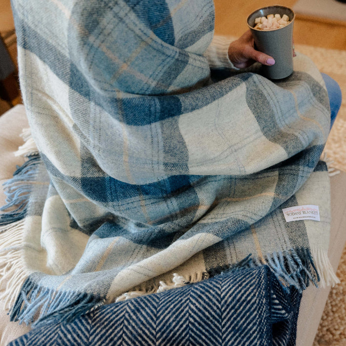 Person wrapped in a Blue Skye Check Shetland Wool Blanket, a blue and white plaid throw, holding a mug of hot chocolate in a living room
