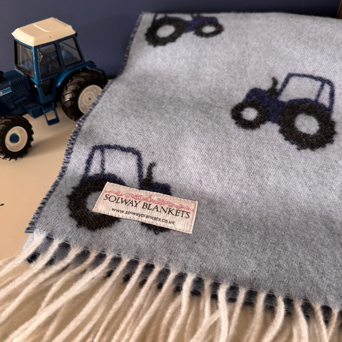 A light blue soft and cosy woven baby blanket with dark navy tractor motifs and cream fringe, laid on a tabletop next to a blue toy tractor. A sewn label reads Solway Blankets.
