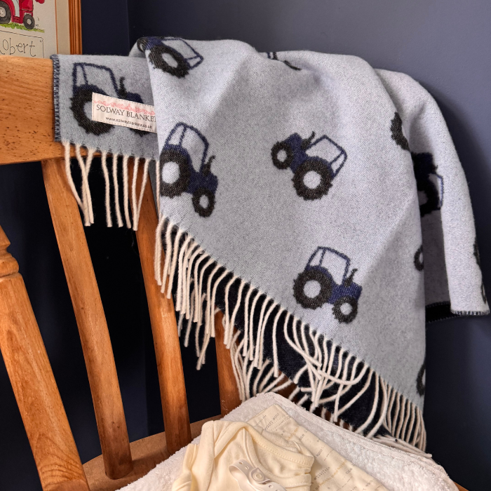 A light blue woven tractor merino lambswool baby blanket with dark blue tractor motifs and cream fringe, draped over a wooden chair in an indoor setting.