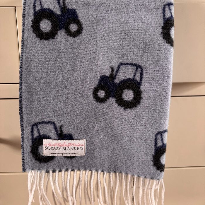 A Blue Tractor Merino Baby Blankets, light blue background with a repeating dark navy tractor pattern and white fringe, featuring a Solway Blankets brand label.